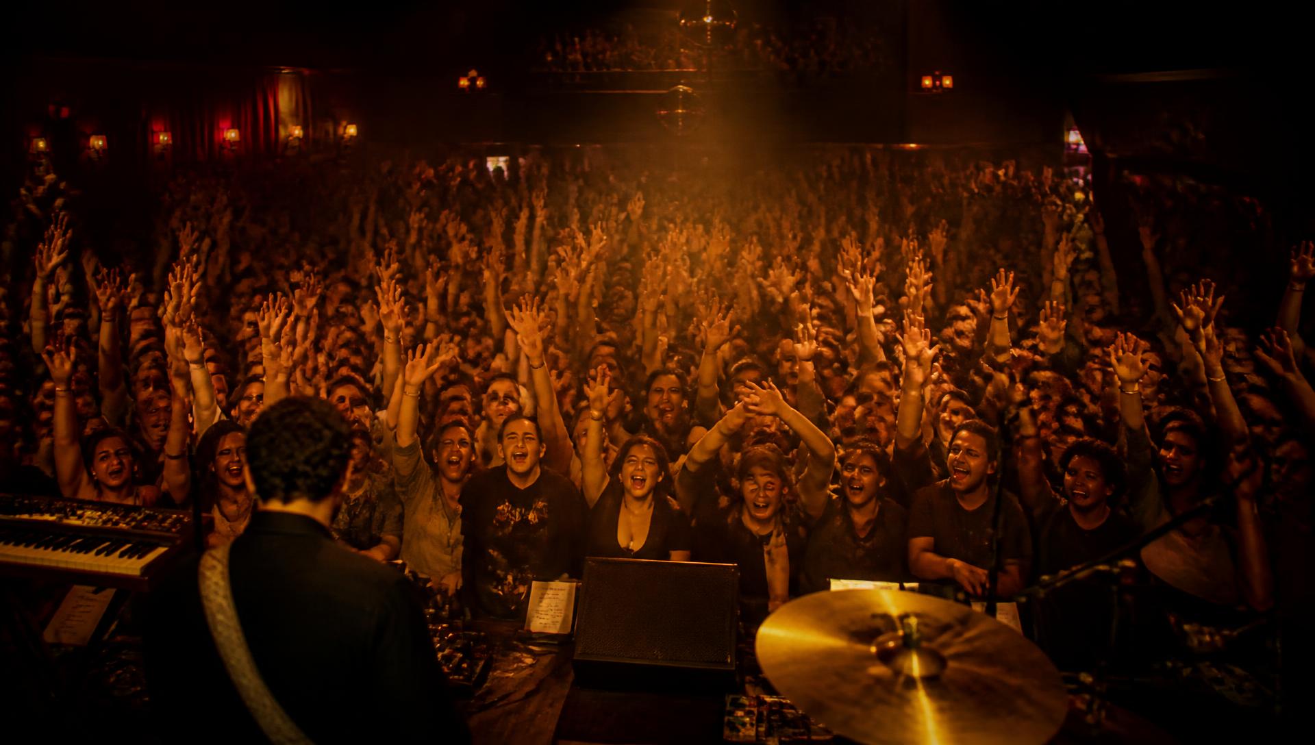 Cheering audience with hands raised in front of a warmly lit concert hall stage